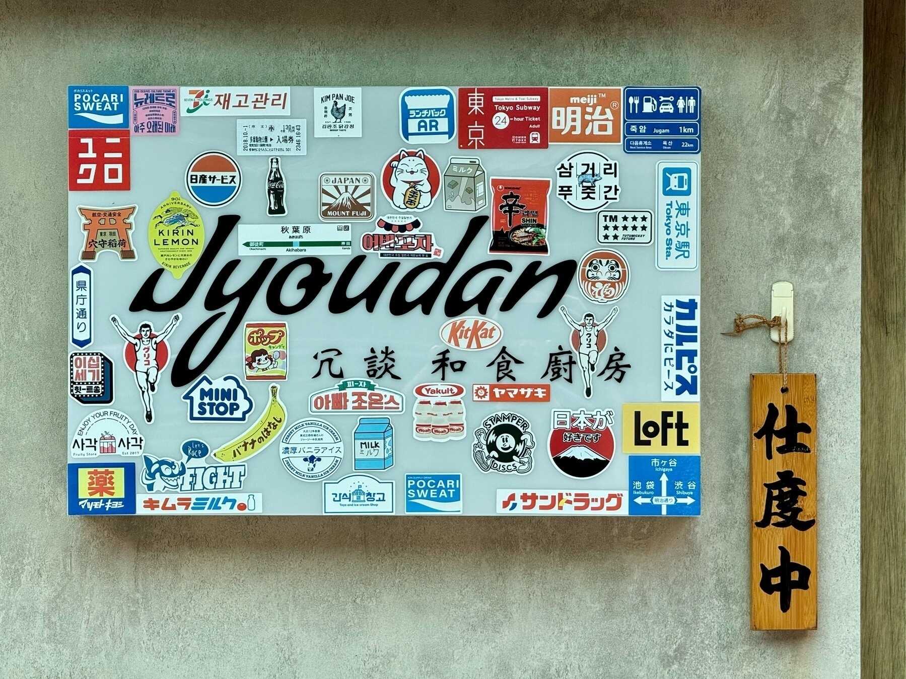 A metallic silver shop sign for a restaurant called 冗談 ("joke"), romanised as "Jyoudan", with lots of different Japanese stickers covering it