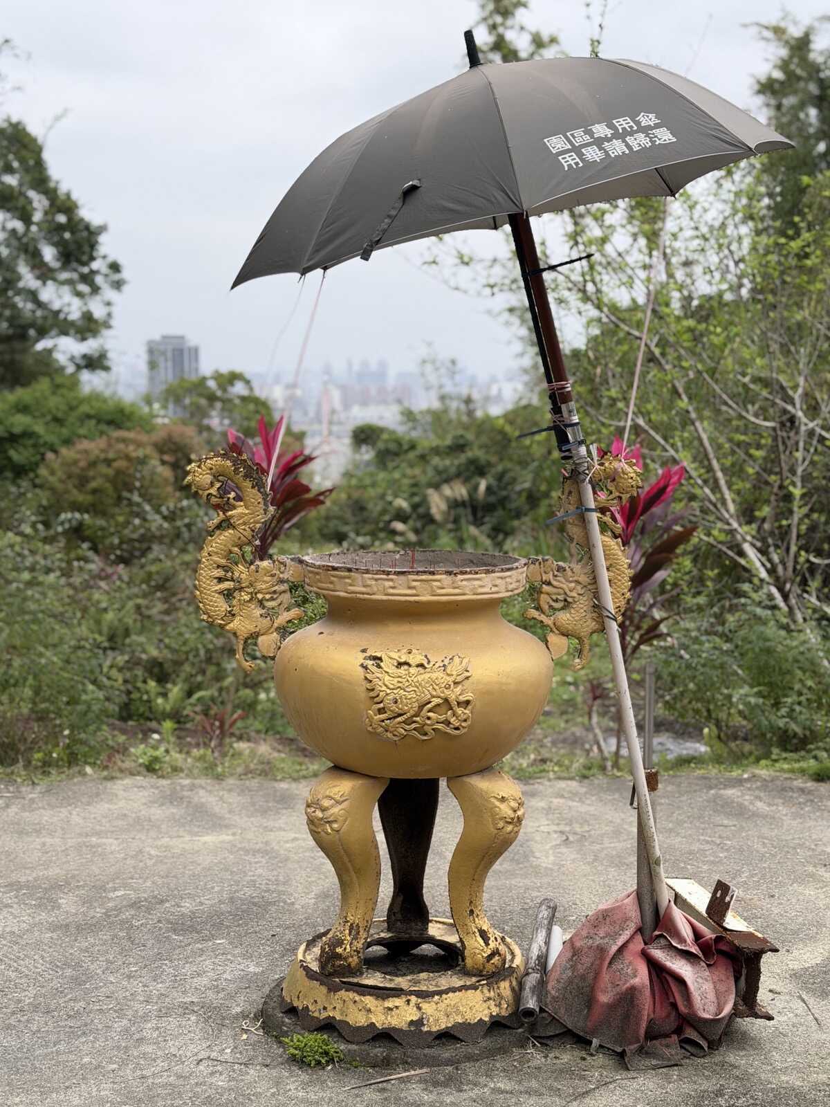 A black umbrella leaning tilted over a temple's large golden incense burner, with a city view in the background. There is mandarin writing on the umbrella